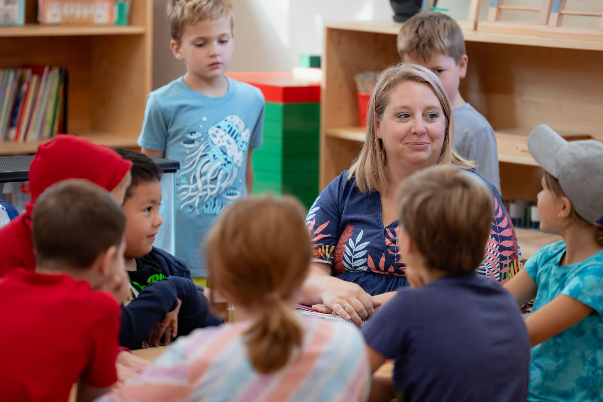 A montessori guide is sitting in a circle with a group of children in a classroom.