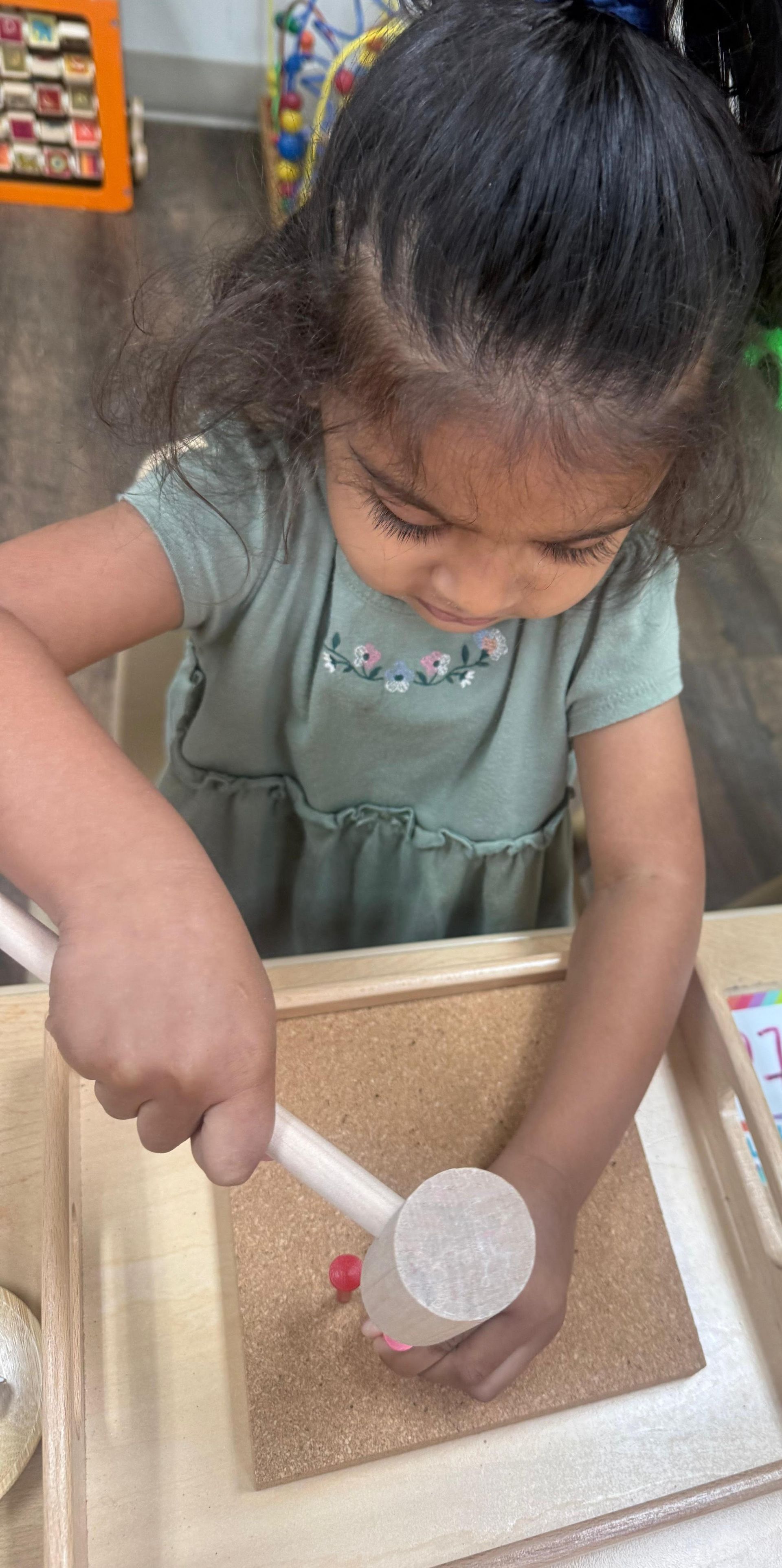 Child using a small hammer while nailing pins into a mini corkboard indoors