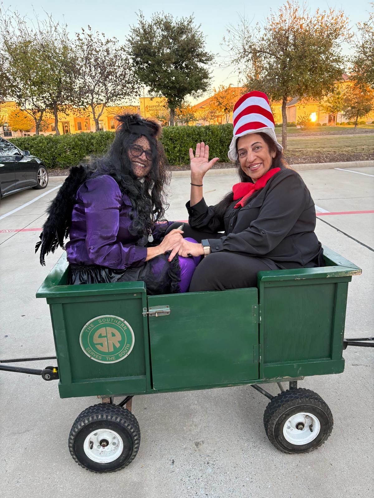 Two people in a green wagon, waving in a parking lot, one wearing a red-and-white striped hat.