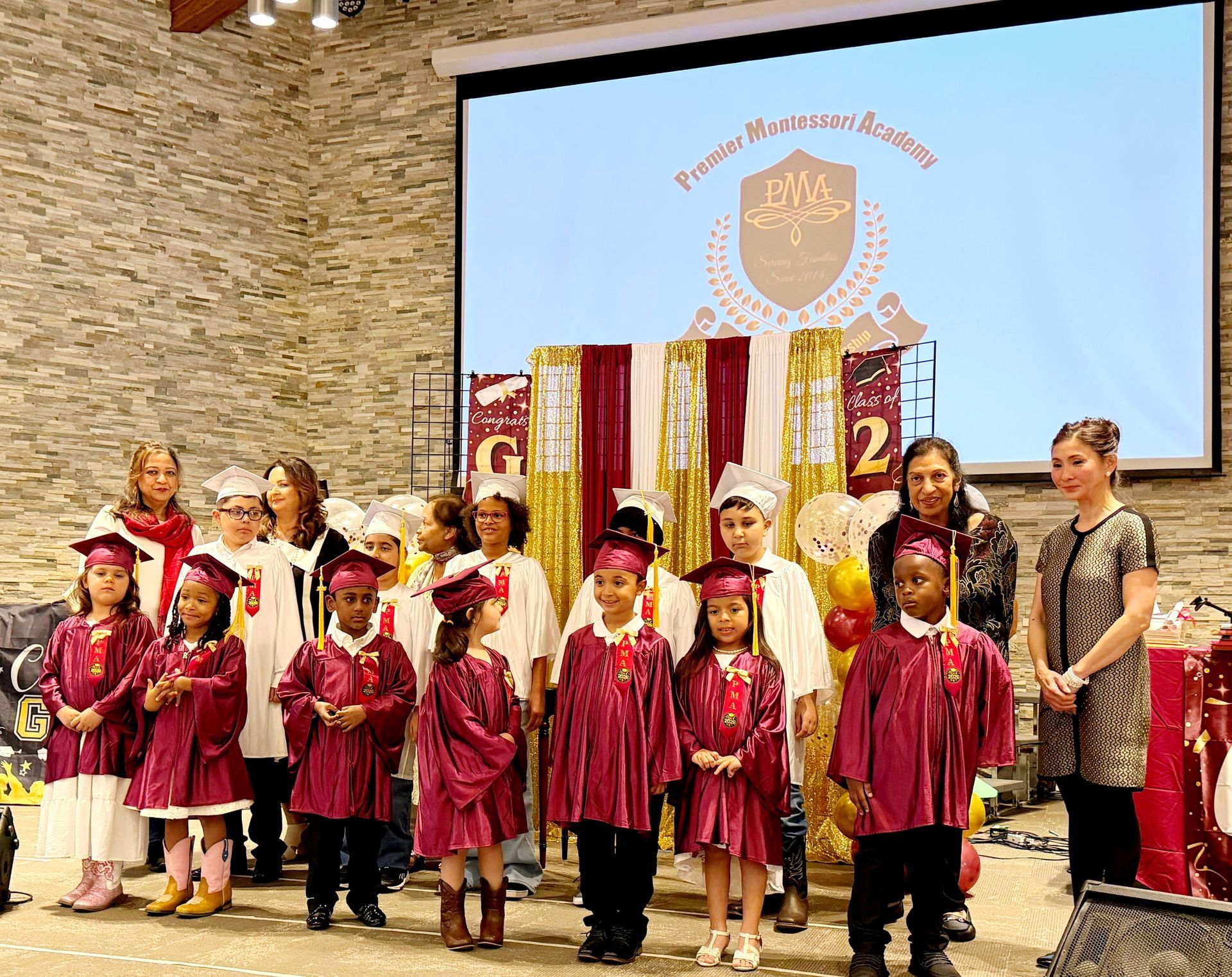 Children in maroon graduation gowns standing on a stage with a school banner and stone wall backdrop