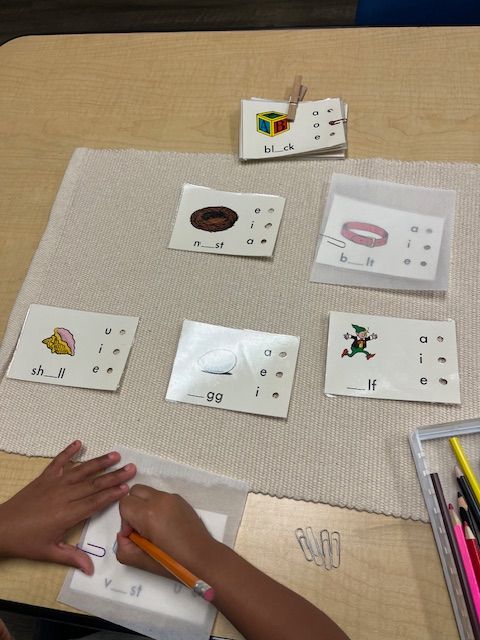 A primary Montessori child practices vowel sounds and phonics by tracing and completing word cards on a work rug.