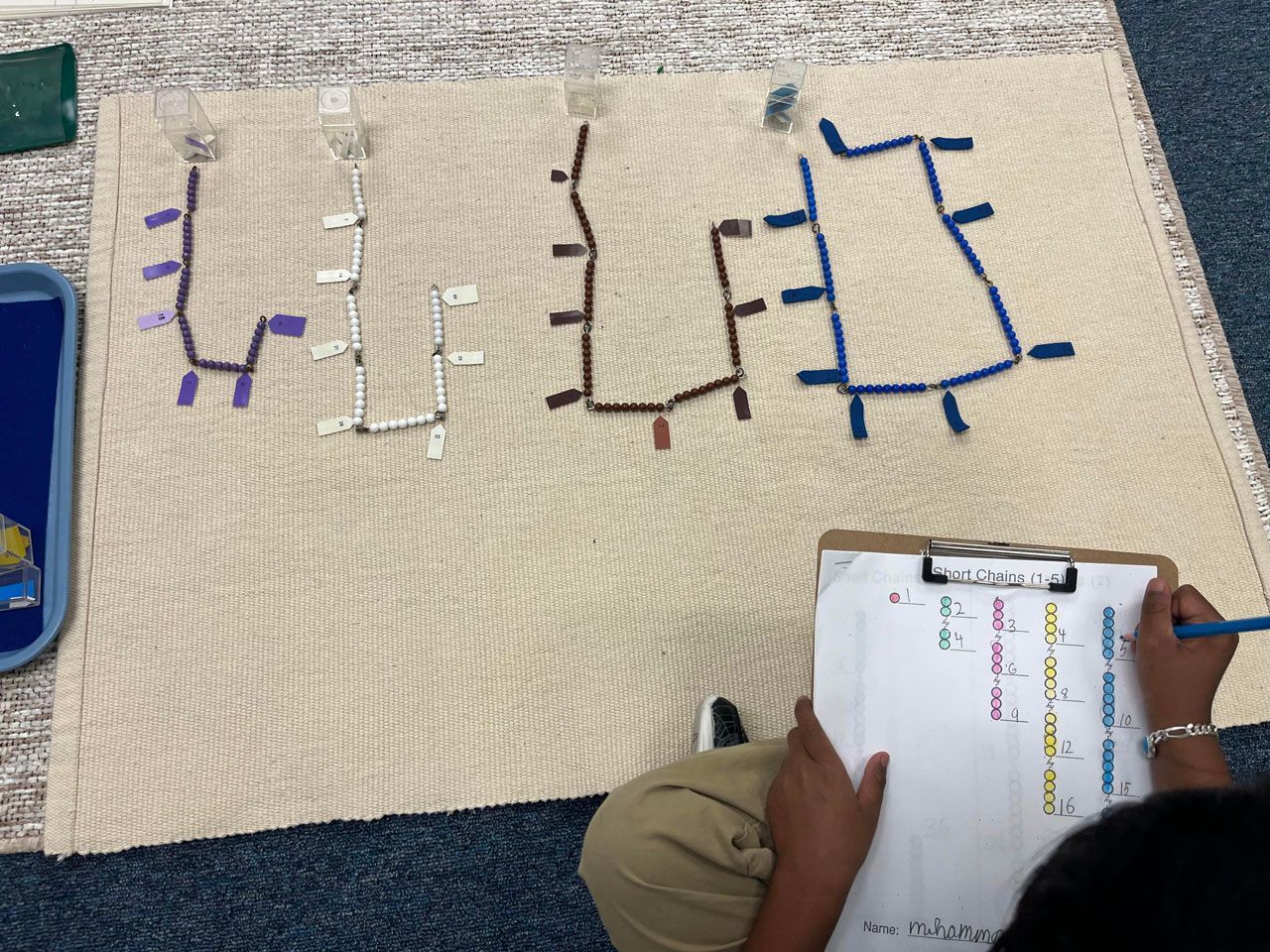 A primary Montessori child uses colorful bead chains and number tickets on a work rug to practice skip counting and linear counting.