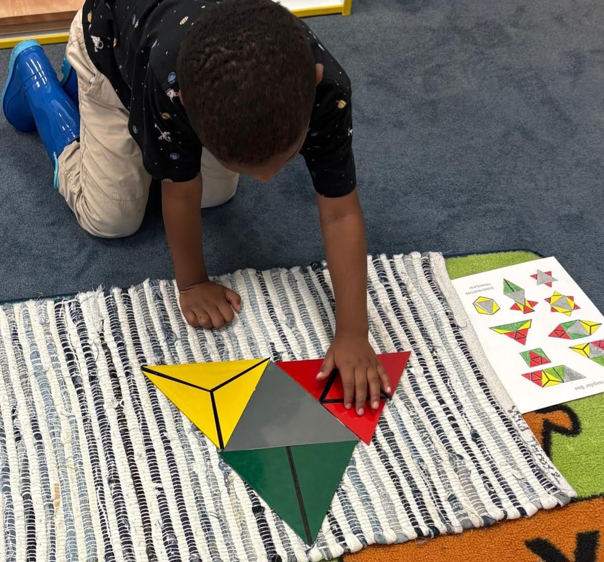 A primary Montessori child uses the constructive triangles material on a small rug to explore how different geometric shapes can form a larger triangle.