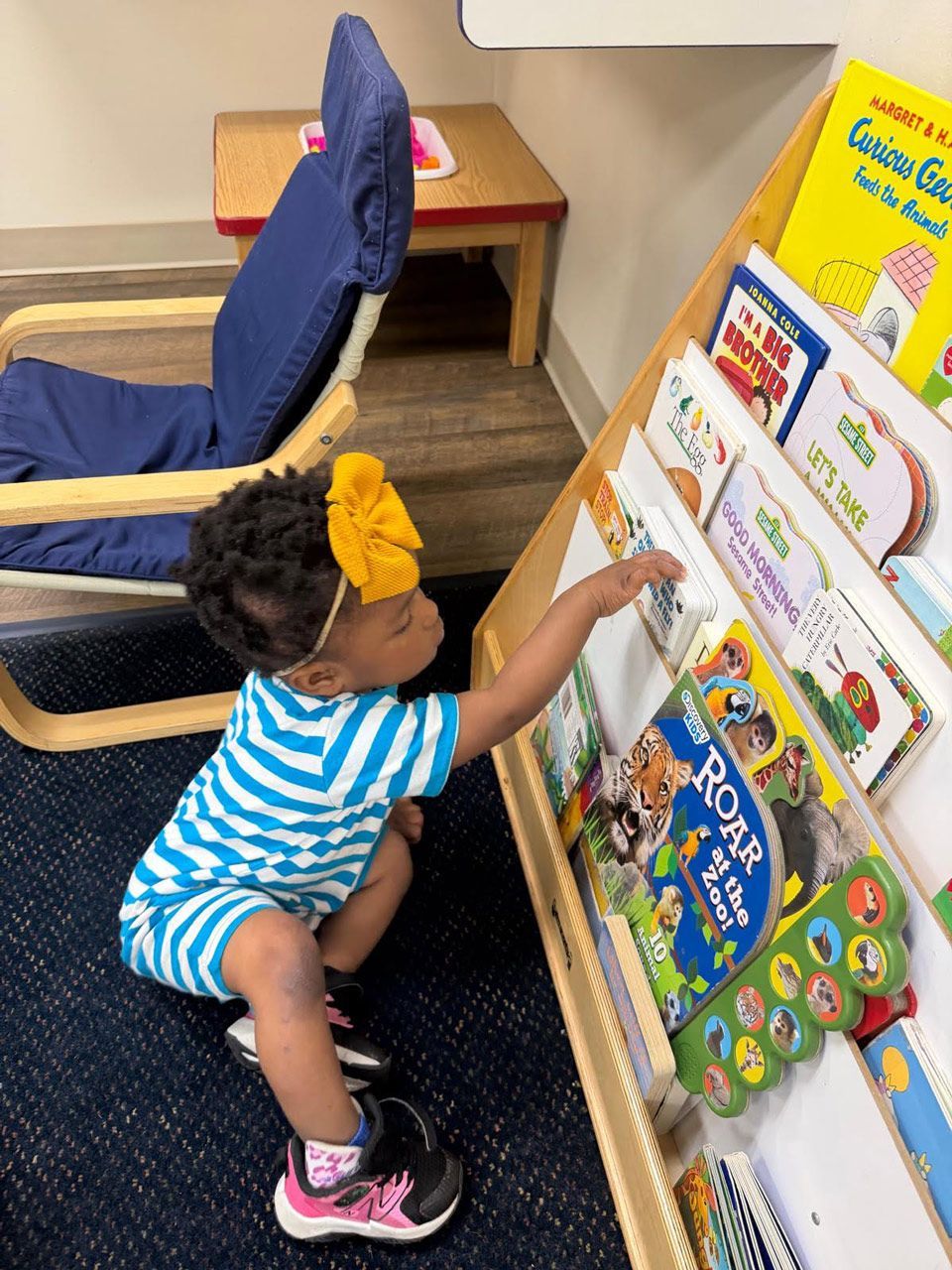 A toddler Montessori child independently chooses a board book from a low, forward-facing library shelf.