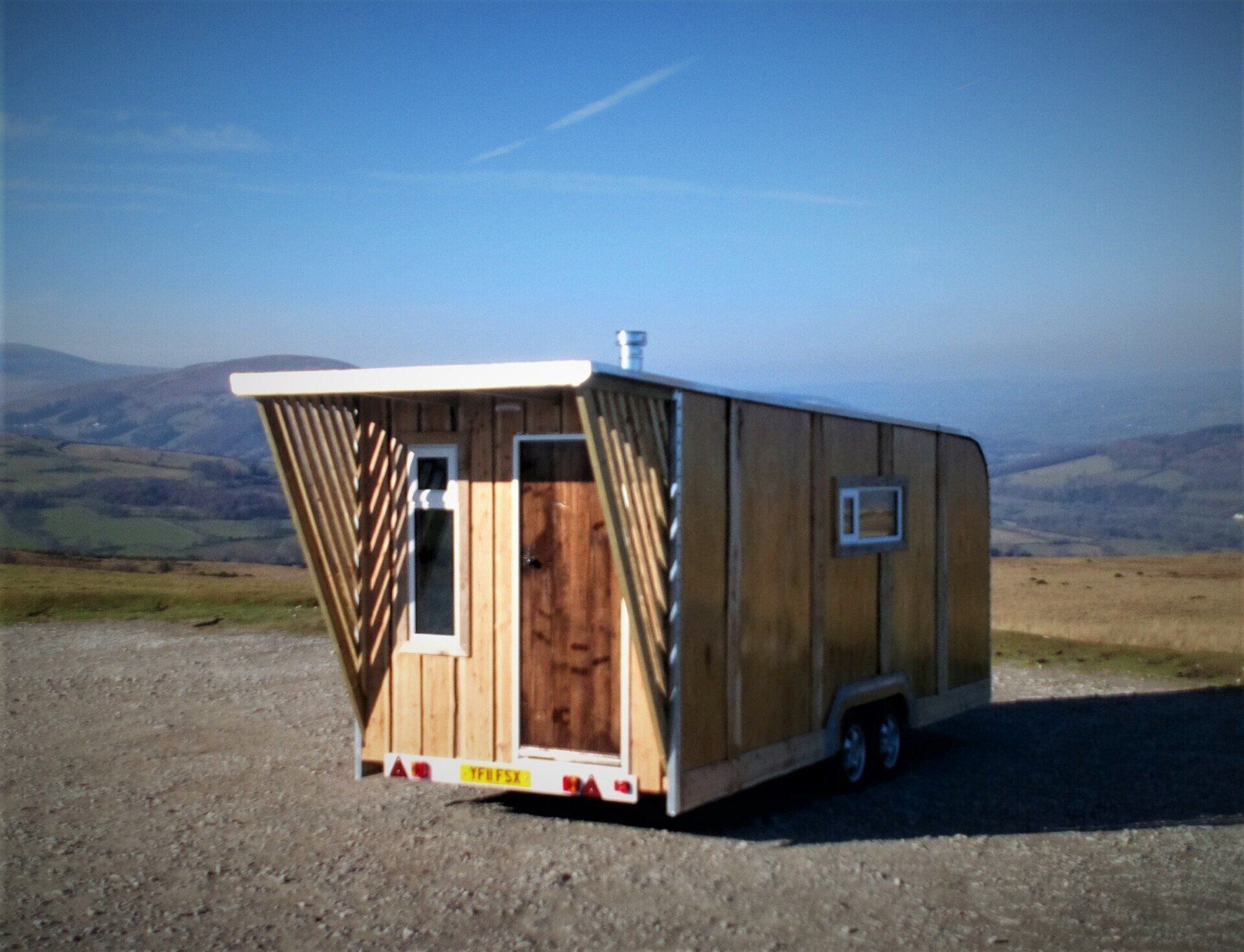 A small wooden house on wheels is parked in a dirt field.