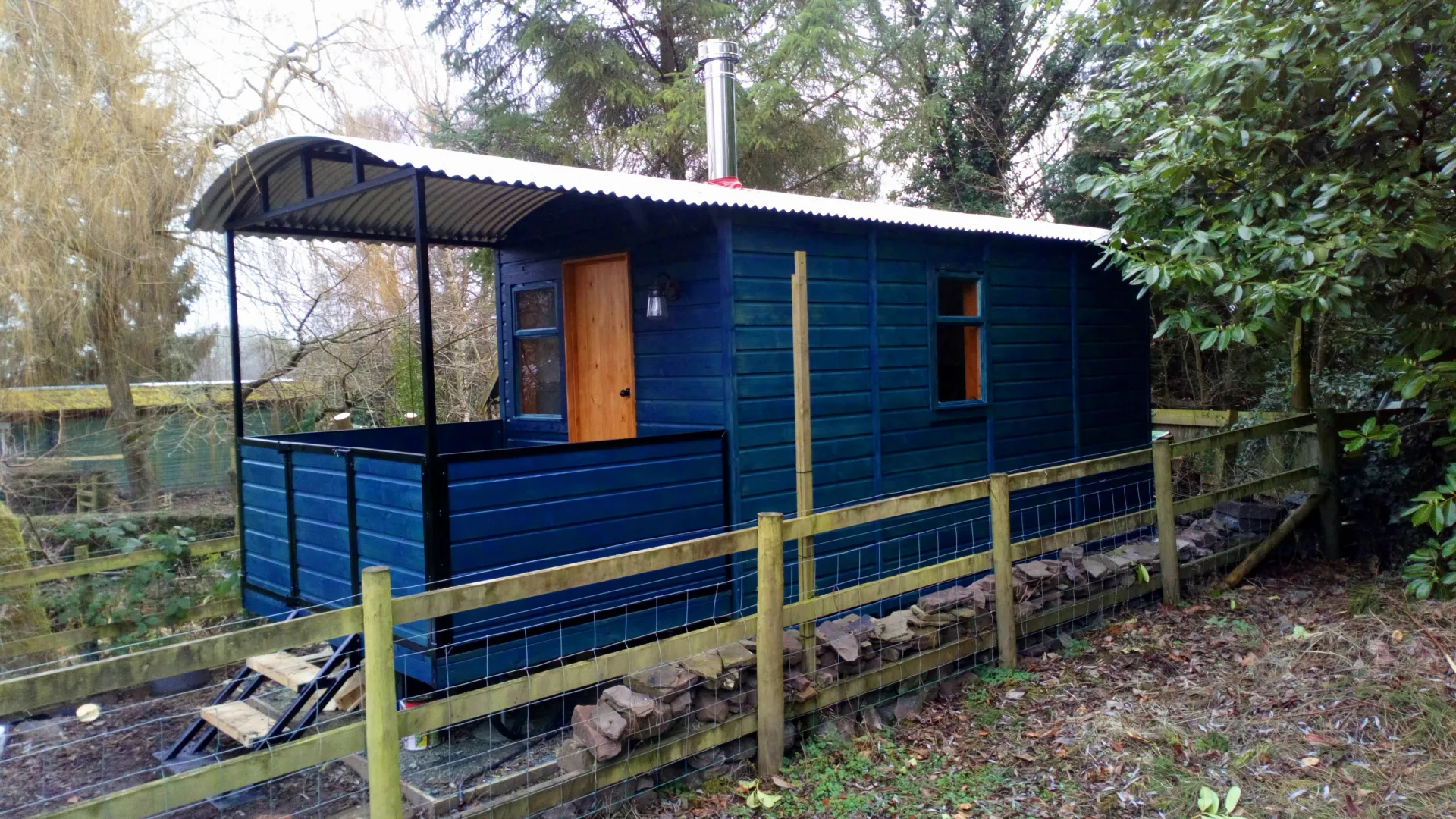 A blue house is behind a wooden fence in the woods.