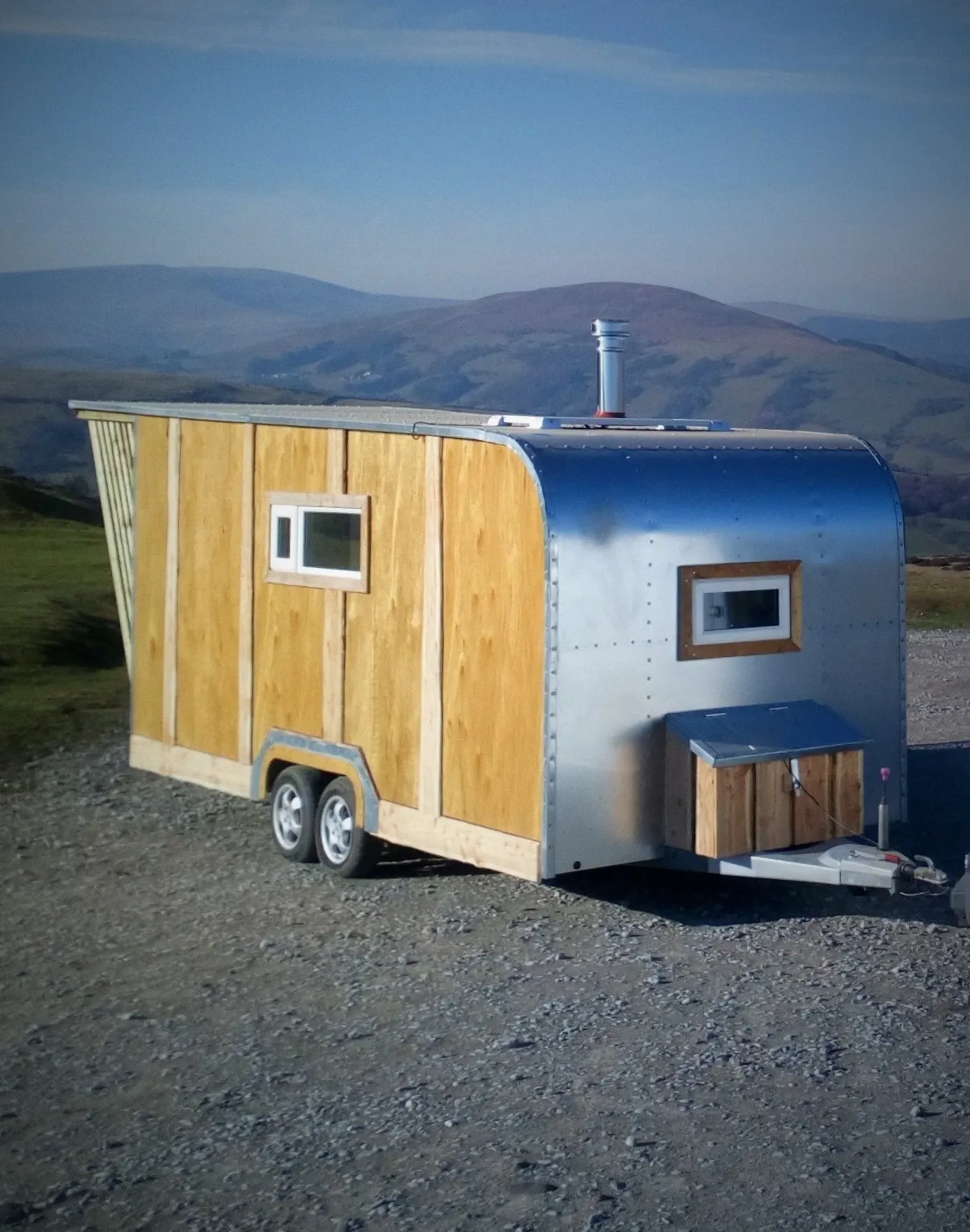 A trailer with a wooden roof is parked on a gravel road