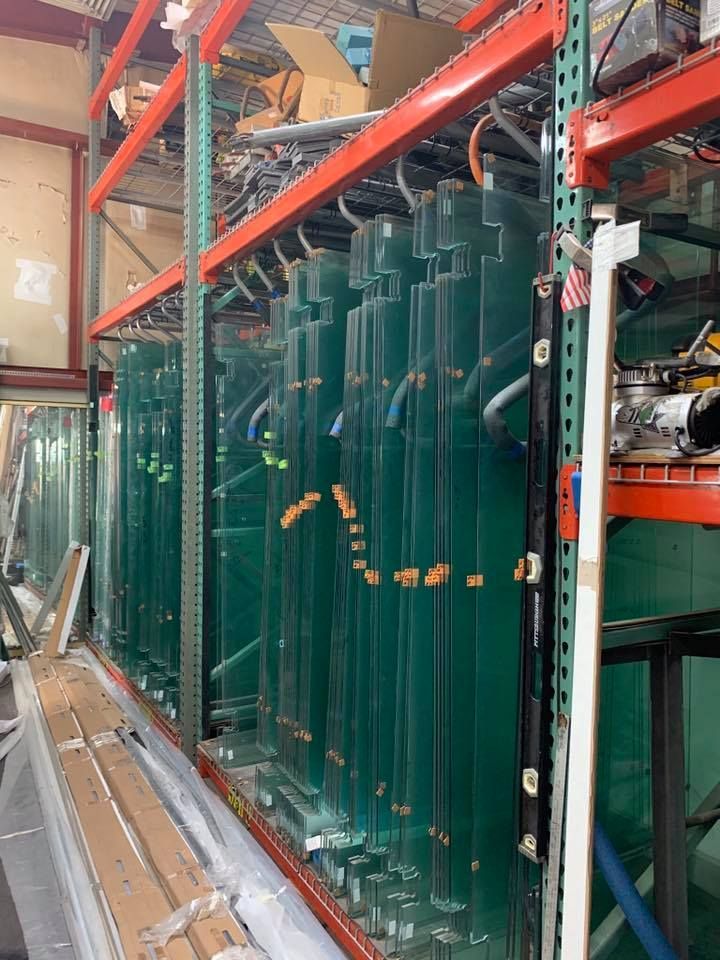 Tall glass panels stored on warehouse shelves, green tinted, with pipes and fittings attached.