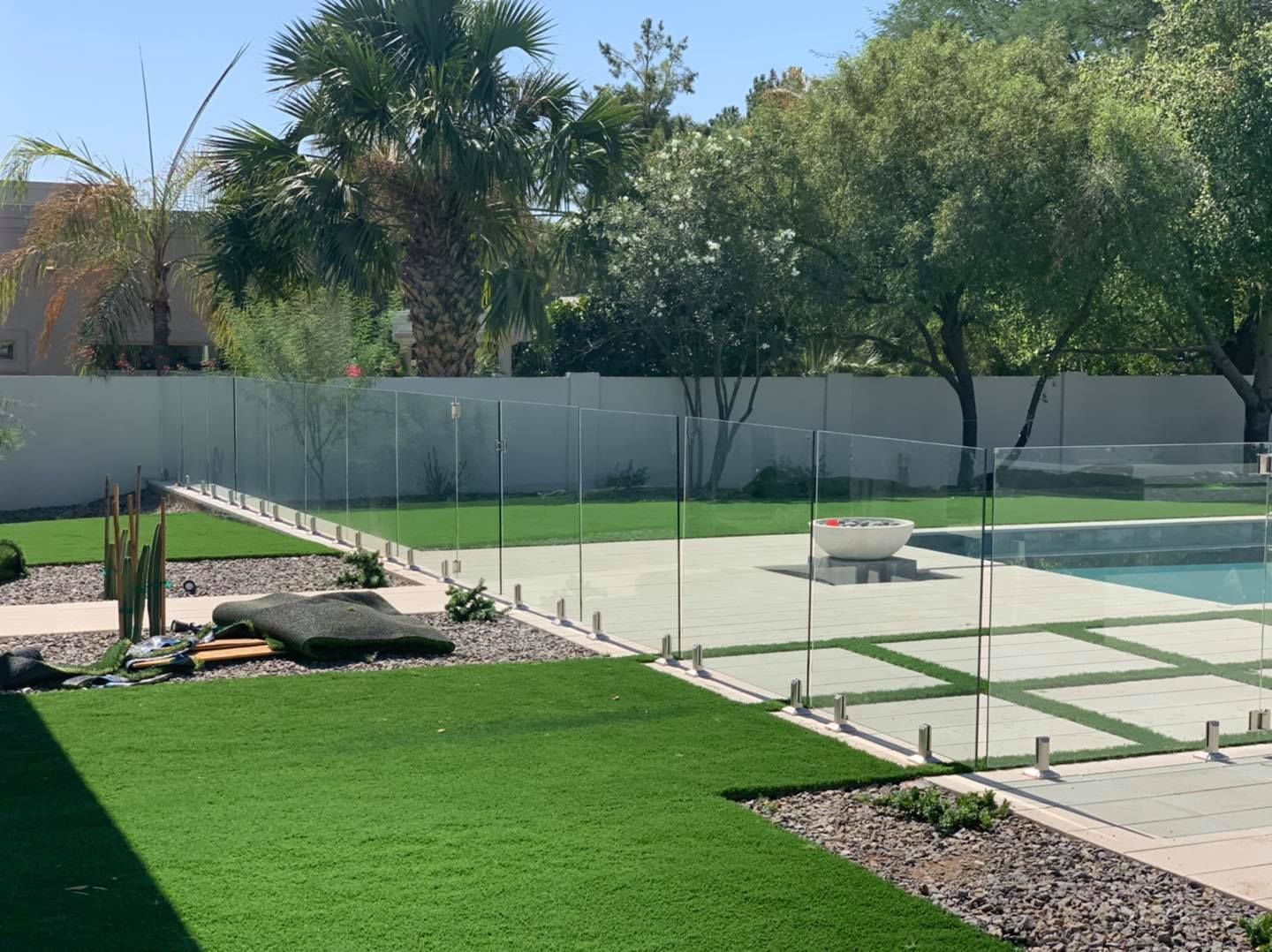A backyard with a clear glass fence surrounding a pool, green grass and plants under a sunny sky.