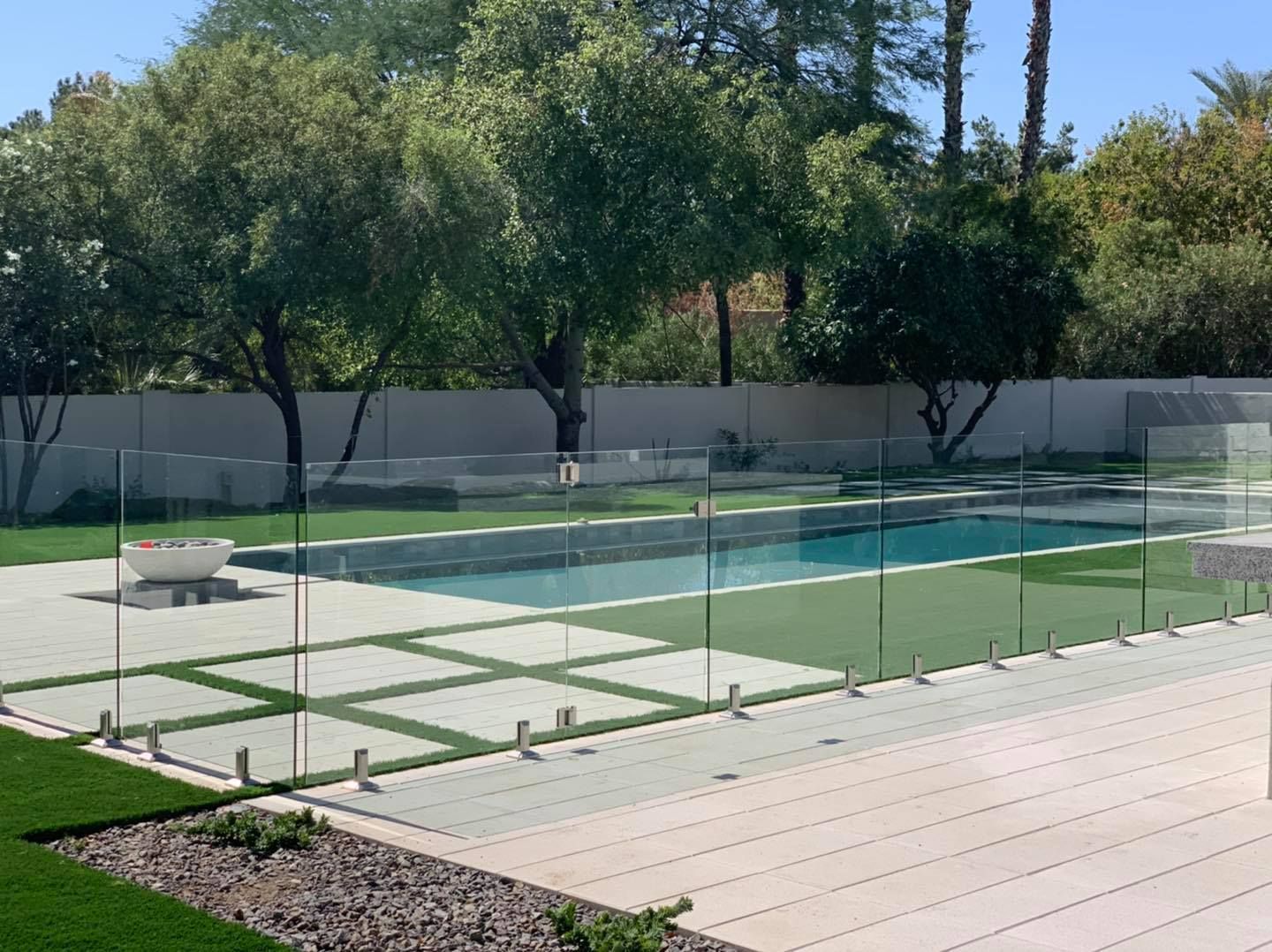 Glass pool fence in backyard with green grass, trees, and a white fire pit.