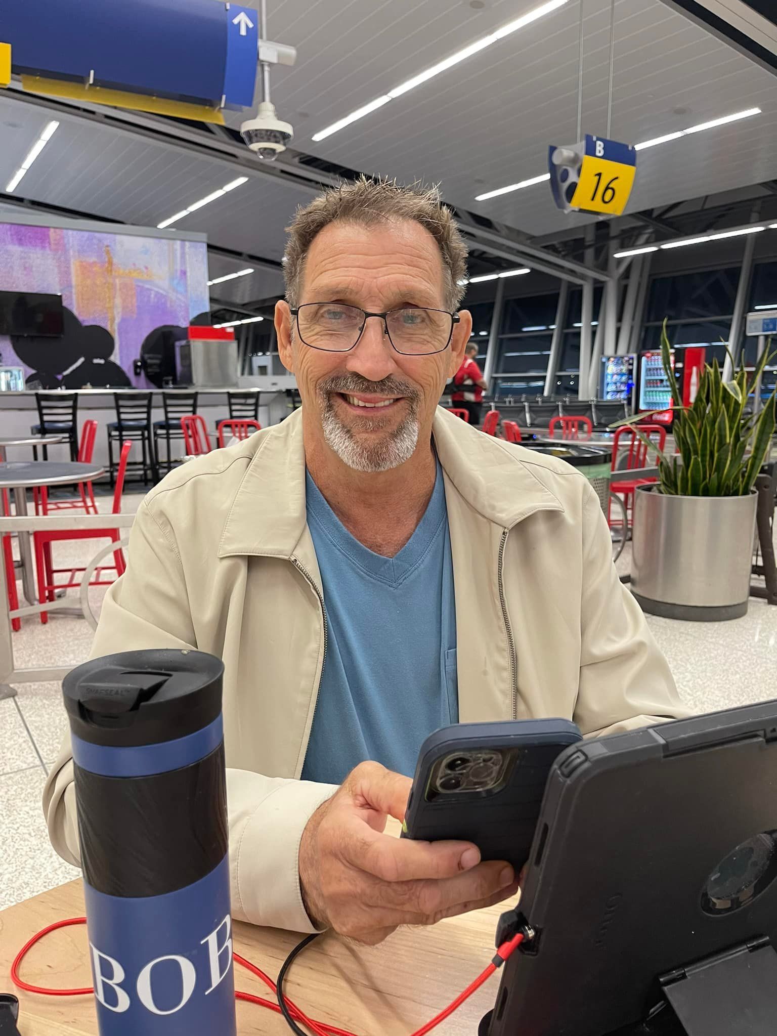 Man wearing glasses at an airport, smiling while using a phone and tablet.