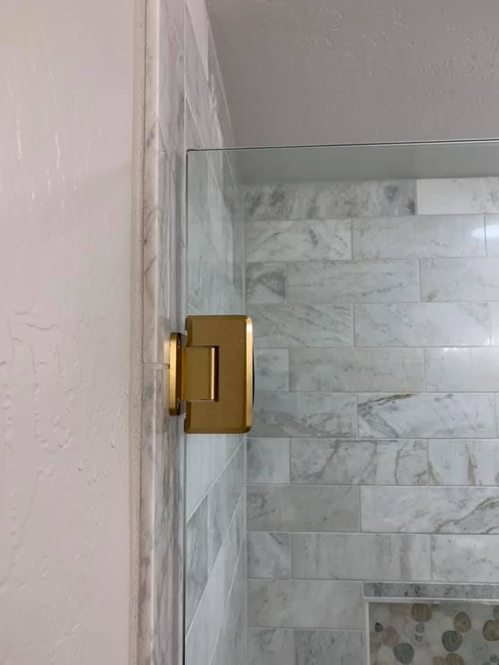 Gold hinge attaching a glass shower door to a marble tile wall.