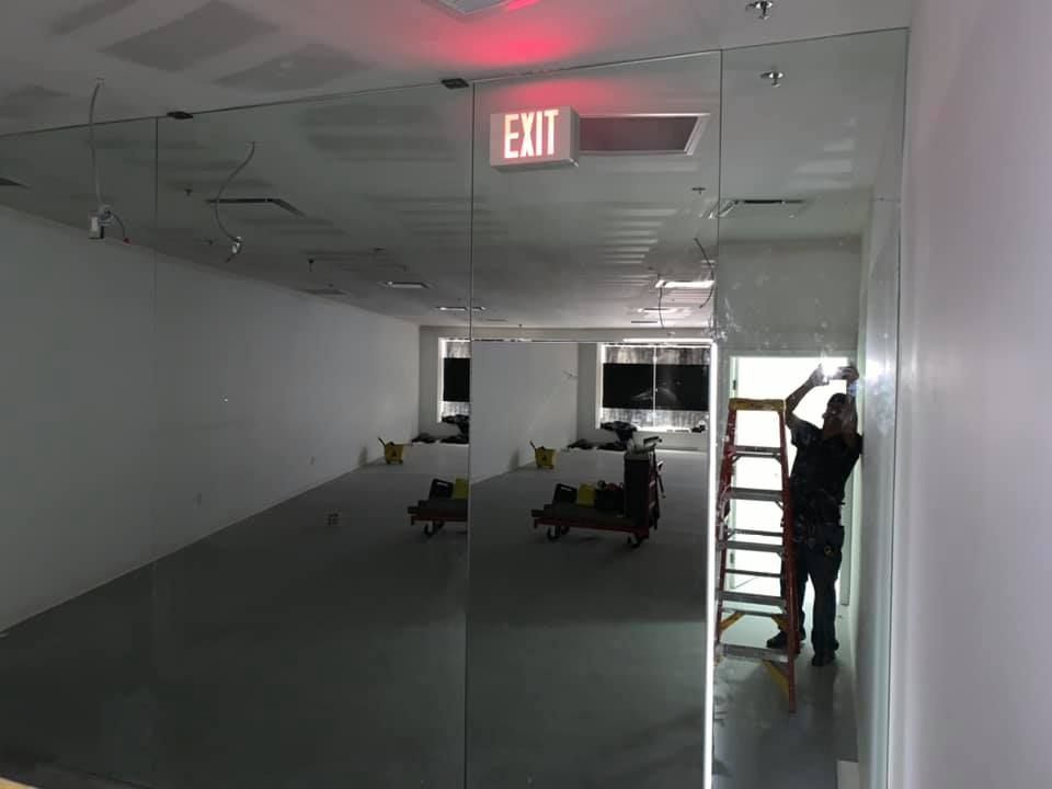 Person installing mirrored wall in a white-walled room, ladder, exit sign above the door.