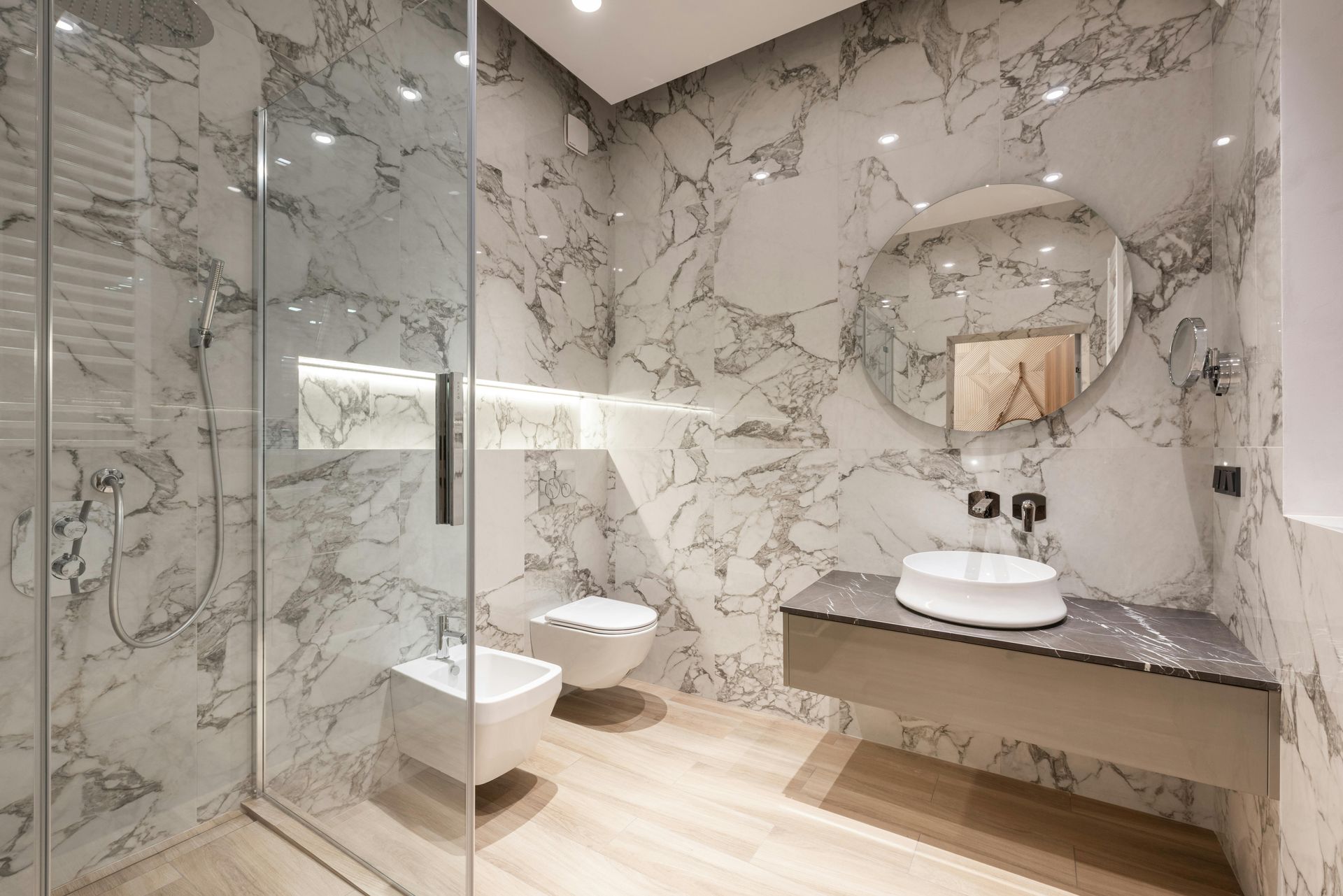 Modern marble bathroom with glass shower, wall-mounted toilet, and sink under a round mirror