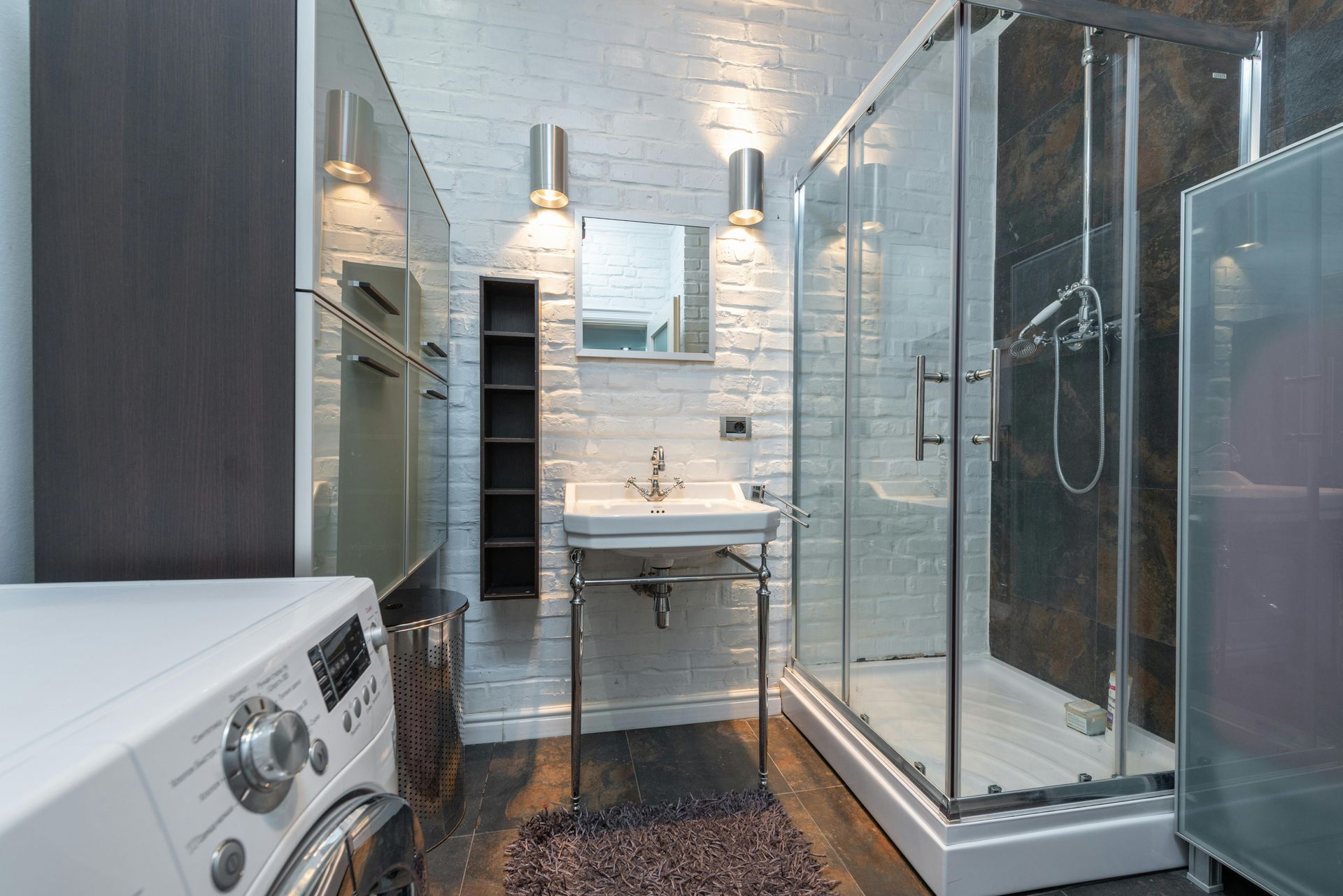 A modern bathroom with white brick walls, a pedestal sink, a glass-enclosed shower, and a washing machine.