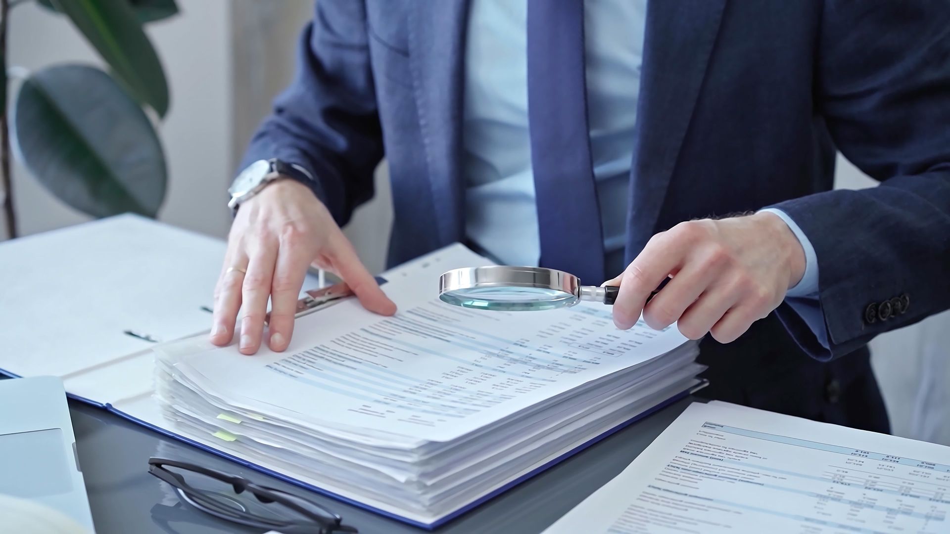 A person in a business suit examines a document with a magnifying glass while sitting at a desk.