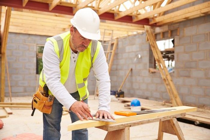 A worker in yellow protective gear and a hard hat is measuring the wood using a ruler.