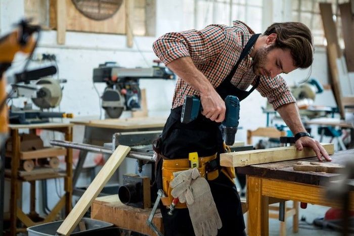 A man with tools on his waist is using a drill to insert the screw on the wood.