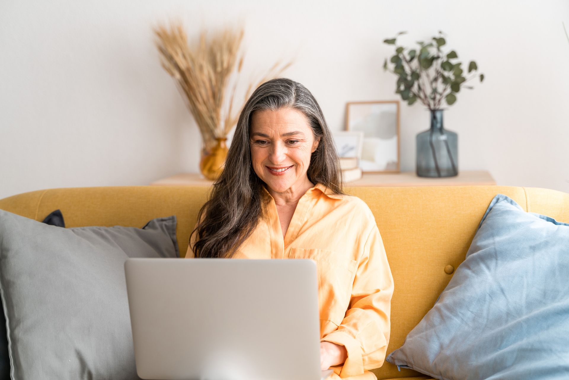 Woman with grey hair, wearing orange shirt, smiles while using a laptop on a yellow couch.