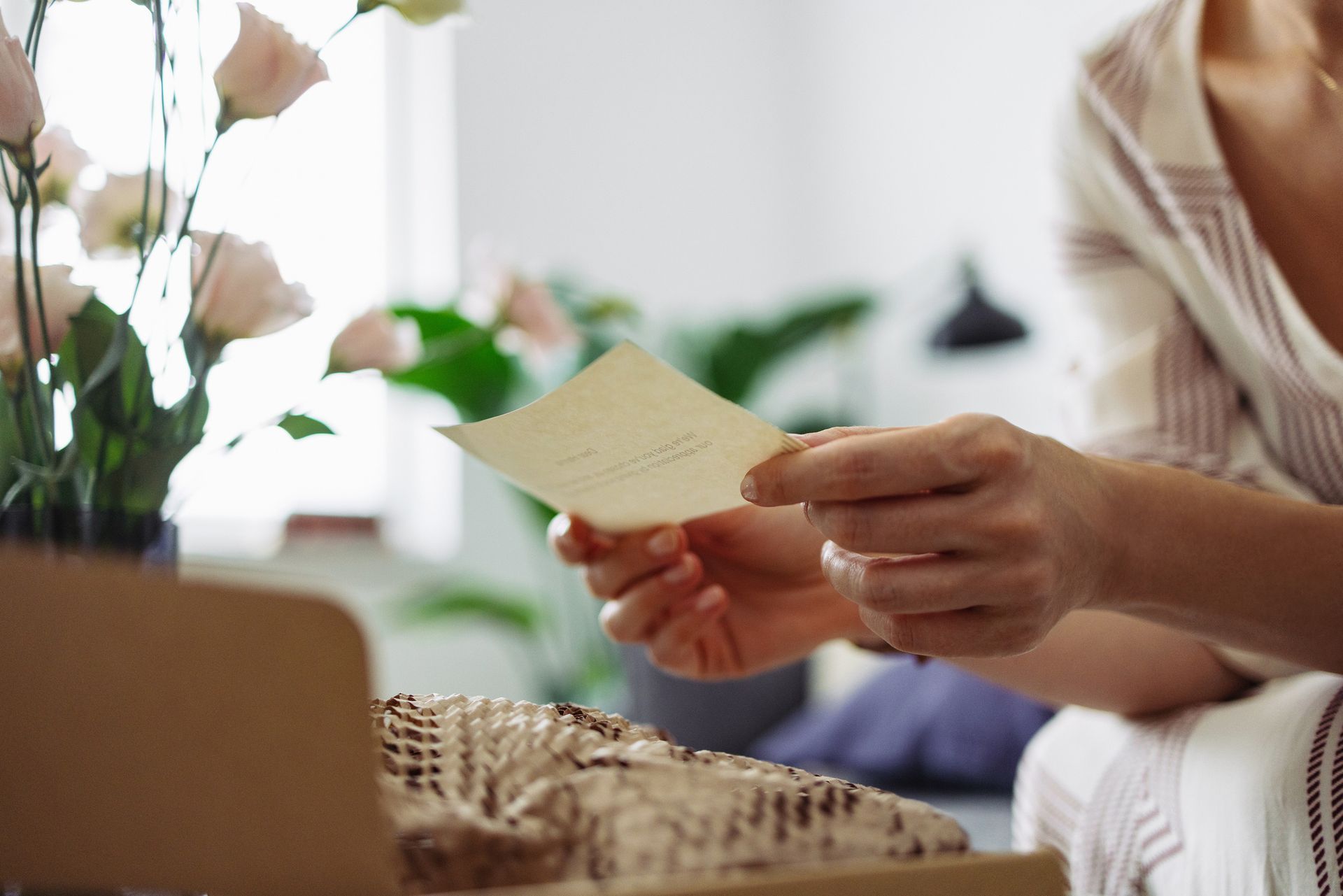 Woman reading a card, holding it in both hands. Flowers and a box sit nearby in a light-filled room.