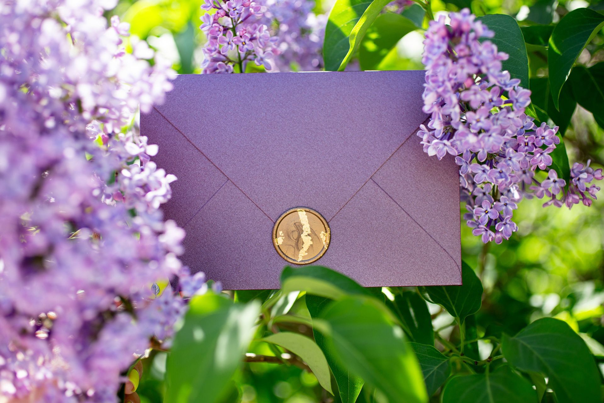 Purple envelope with a gold seal nestled in lilac blossoms.