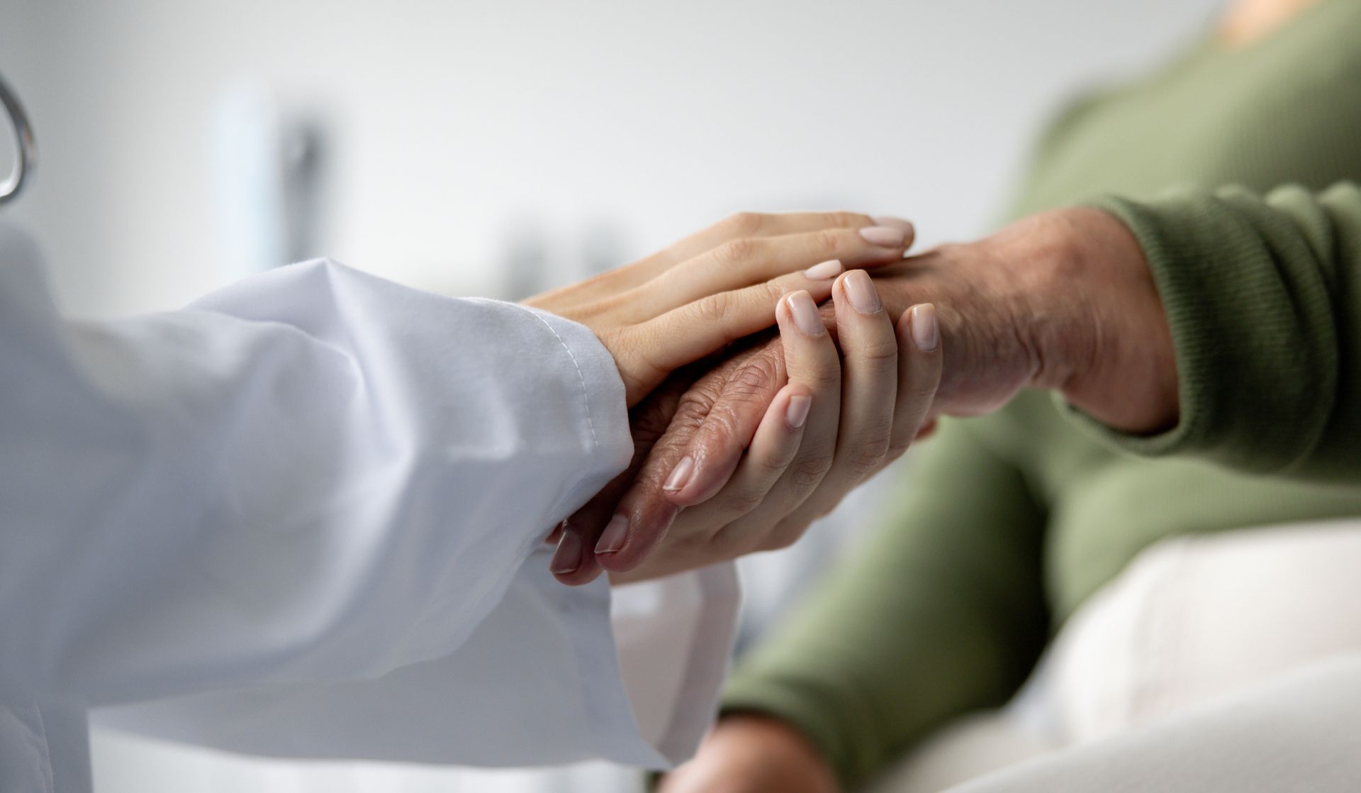 Woman kneeling by lake, releasing something into the water; beige hat, black clothing, overcast sky.