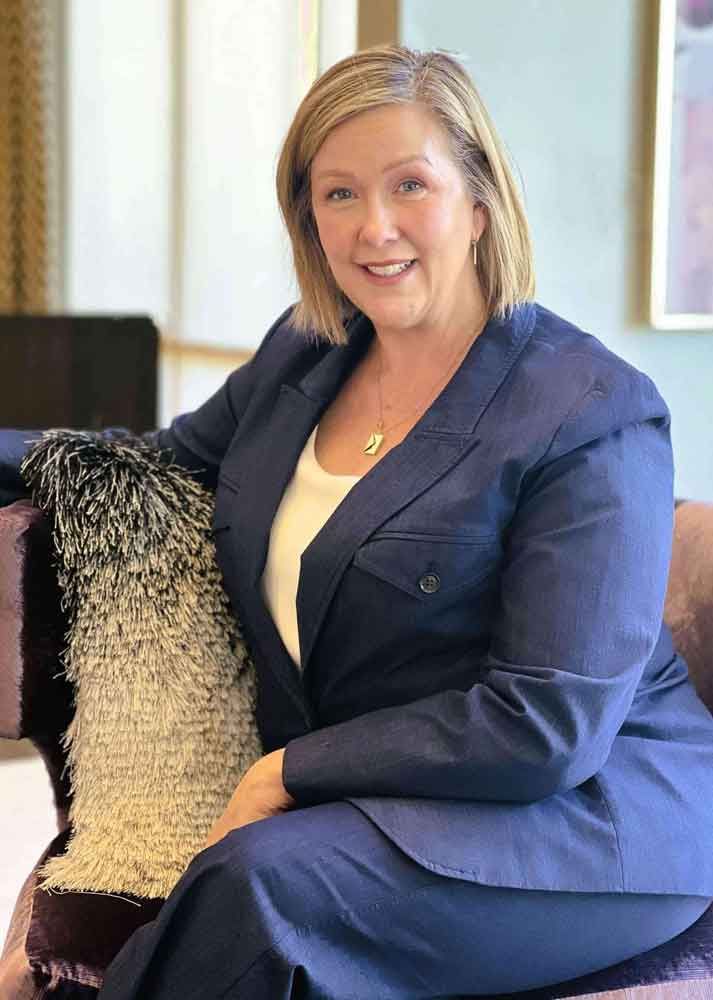 Woman in blue suit seated on a purple couch, smiling.