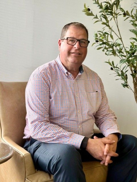 Man seated in chair, wearing glasses and checkered shirt, hands clasped.  Slight smile.  Plant in background.