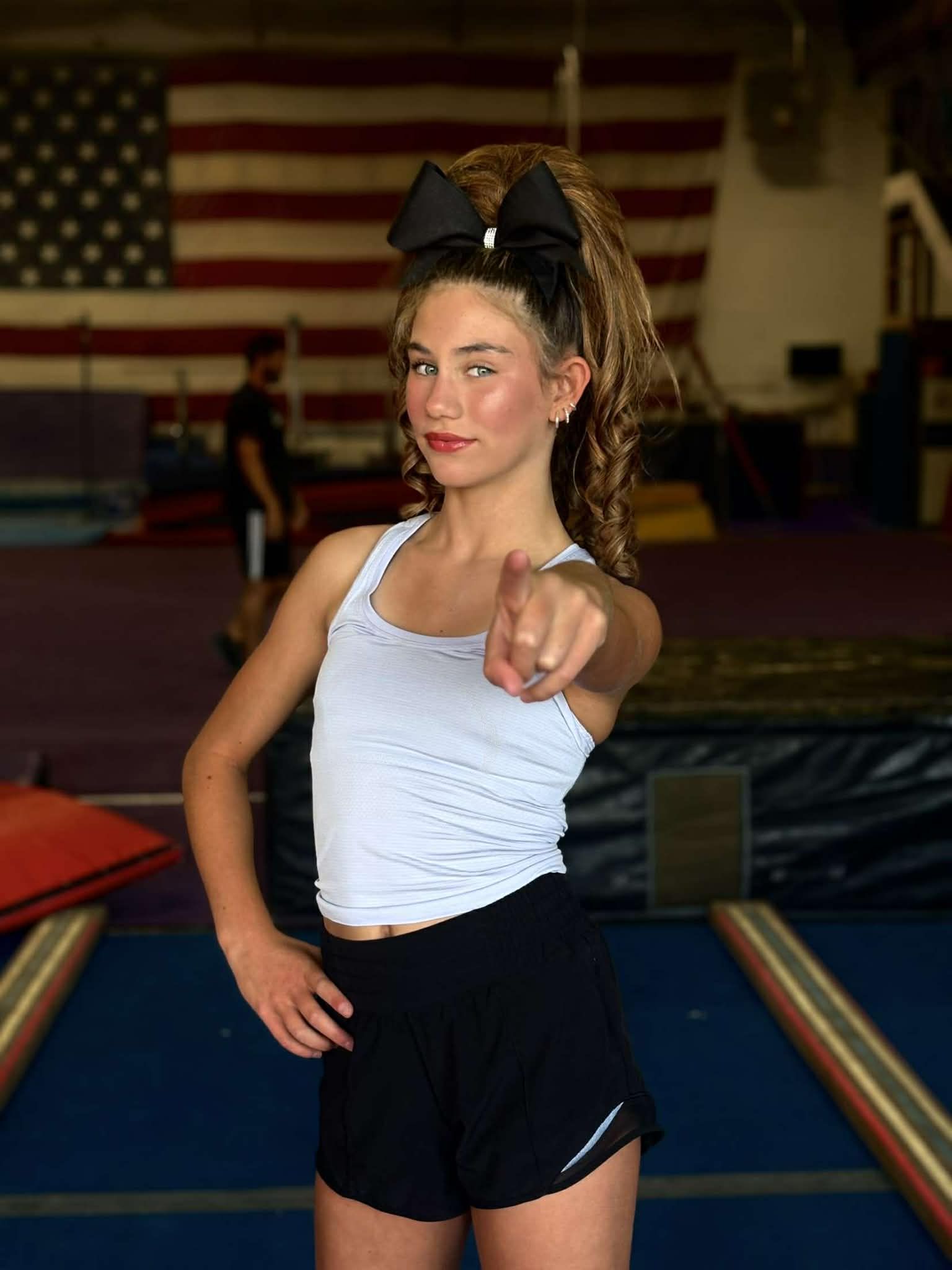 Girl in black and white cheerleading uniform, pom-poms held high, balancing on one foot, smiling.