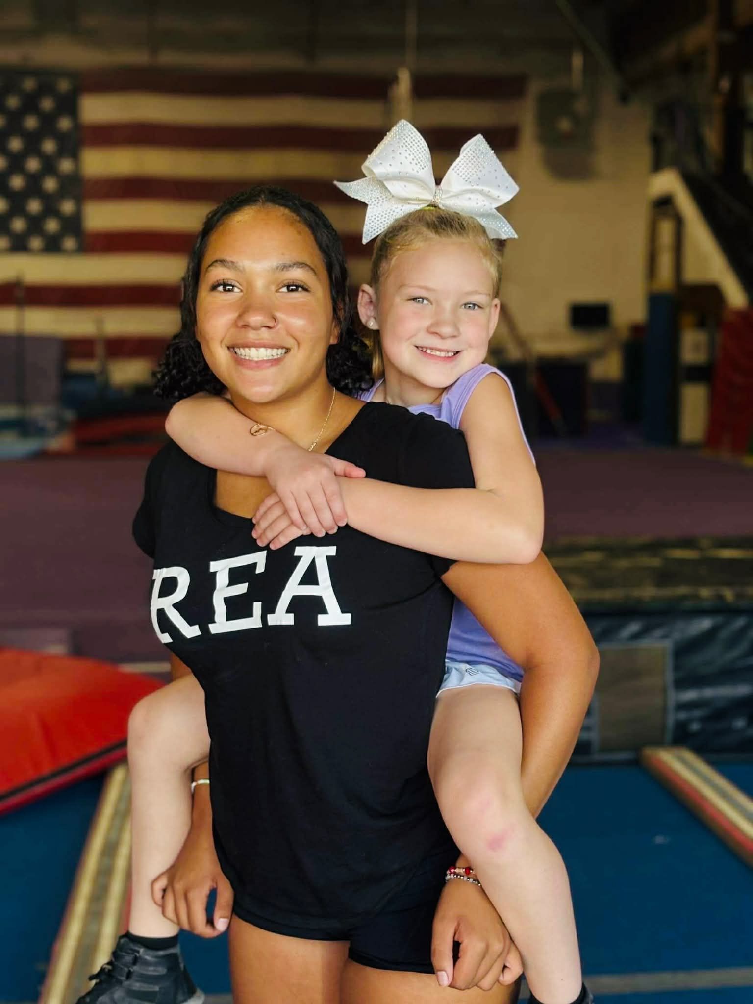 Child sitting on mat, stretching legs, reaching for toes, wearing yellow shirt and purple pants.