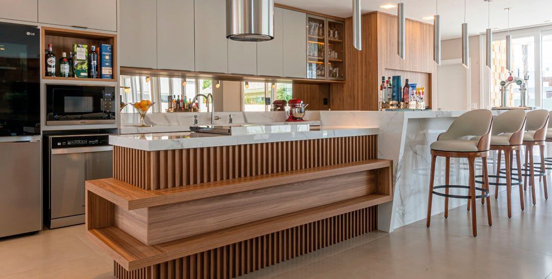 O coração magnético do lar: uma ilha projetada para receber bem e criar conexões. Ampla cozinha planejada com ilha central em madeira ripada, bancada branca e banquetas - Batel Select Curitiba.