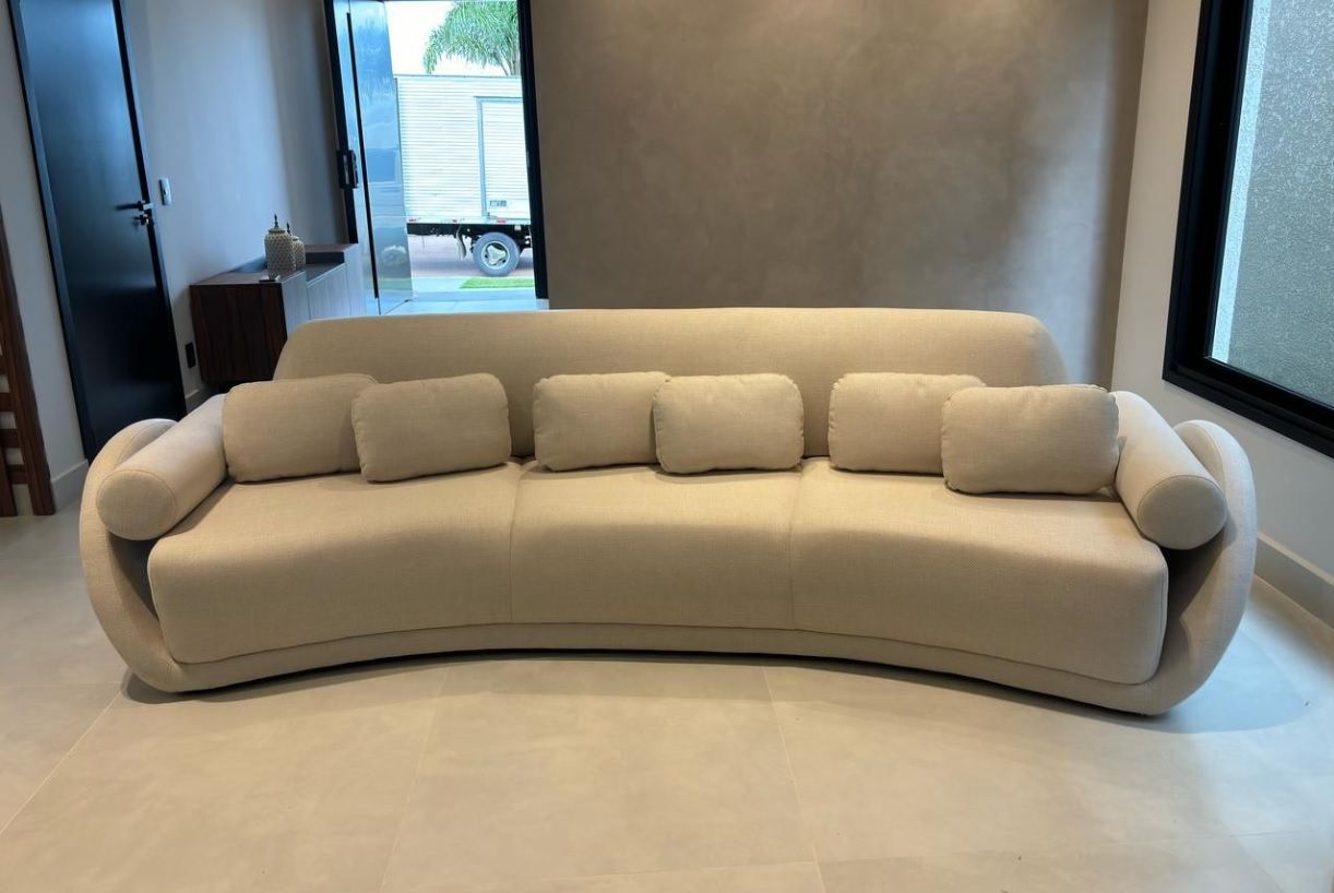 Sofá home de alto padrão em Curitiba, formato curvo orgânico, revestido em tecido bege claro, com almofadas retangulares e braços arredondados, ideal para salas sofisticadas.