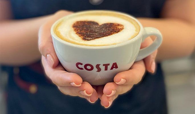 A person is holding a cup of costa coffee with a heart in the foam.
