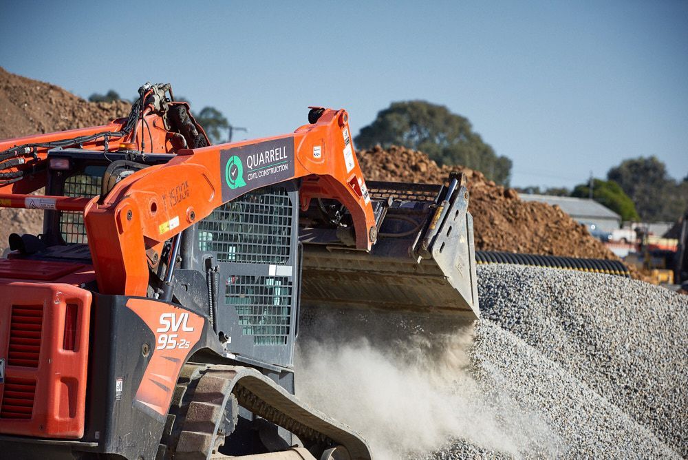 An Orange Kubota Svl 95 Compact Track Loader Scoops Gravel on a Sunny Construction Site — Quarrell Civil Construction In Congupna, VIC