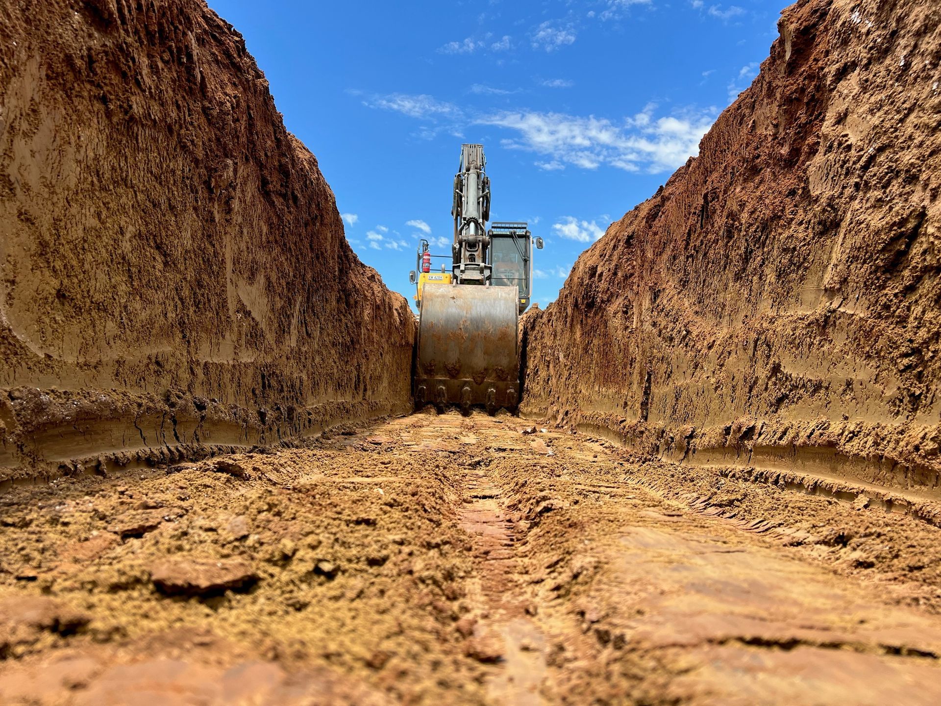 An excavator digging a deep, narrow trench in reddish-brown soil under a bright blue sky — Quarrell Civil Construction In Congupna, VIC
