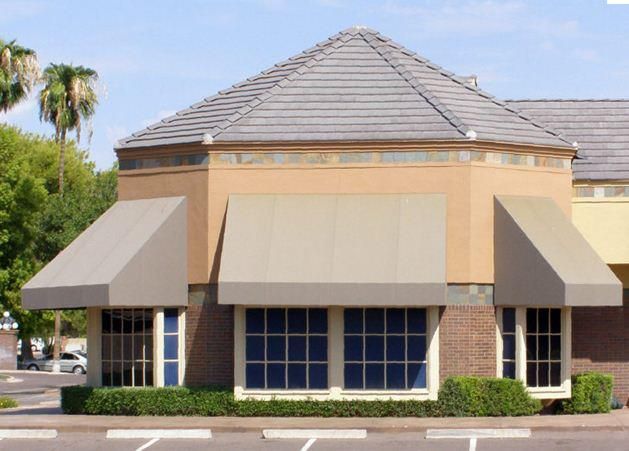 Tan building with angled awnings over windows, tile roof, and low hedge