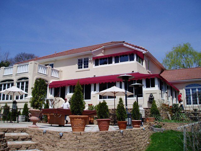 Large, two-story house with a red awning over windows, outdoor patio with tables, pots, and a blue sky
