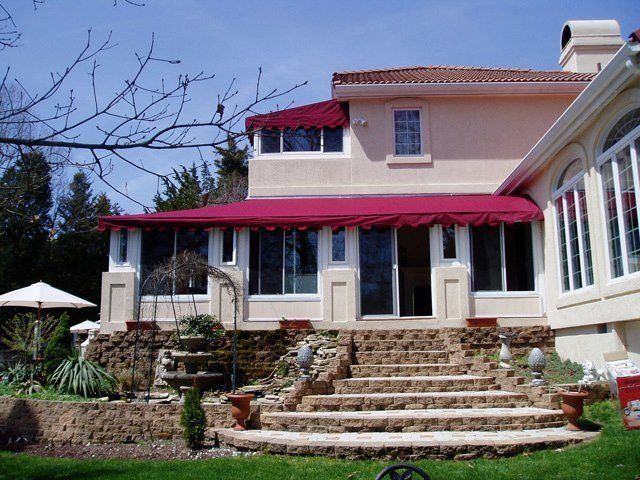 Beige house with burgundy awning over patio, stone steps lead to the door, lawn and fountain in the foreground