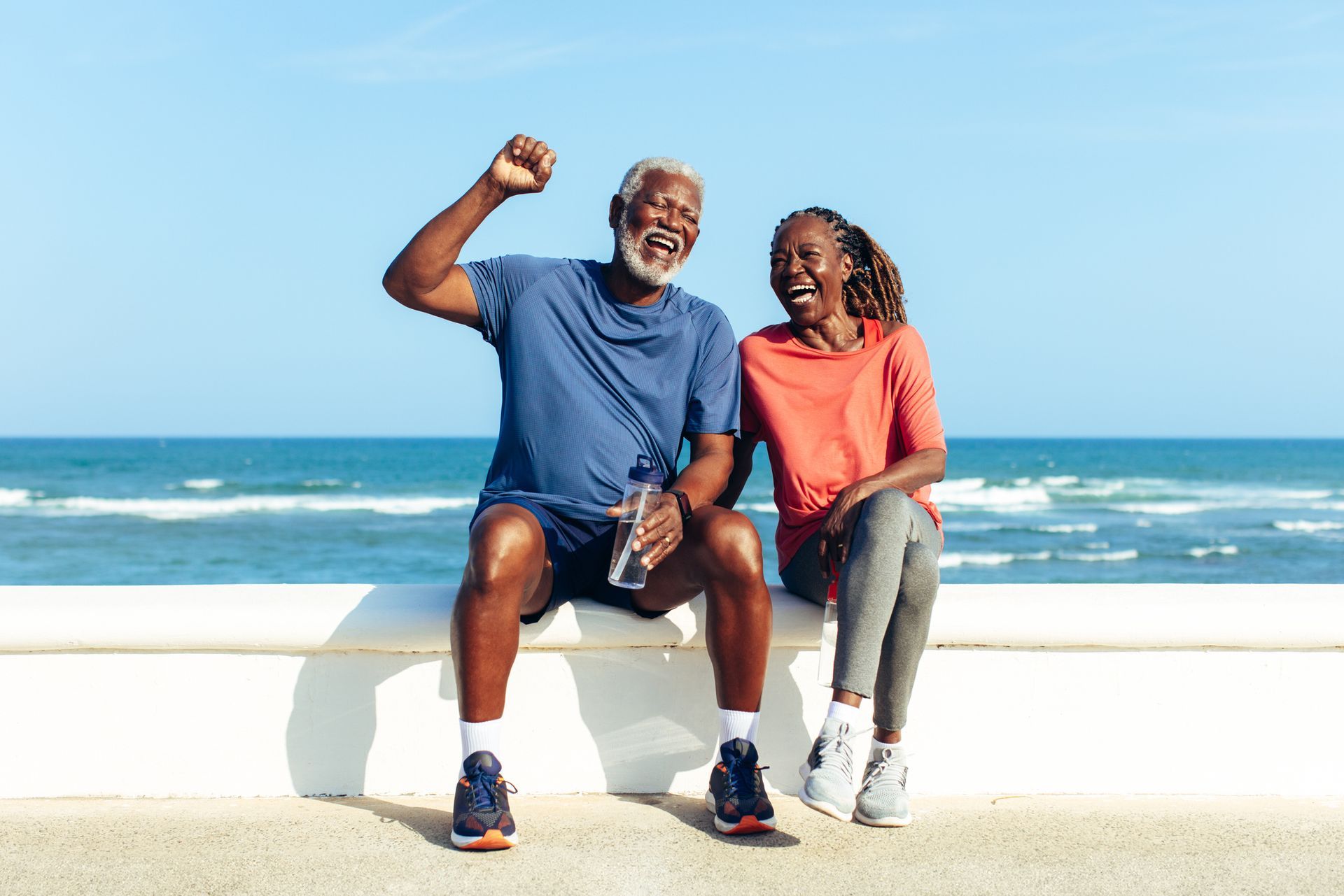 Older couple in activewear sitting by ocean, smiling. 