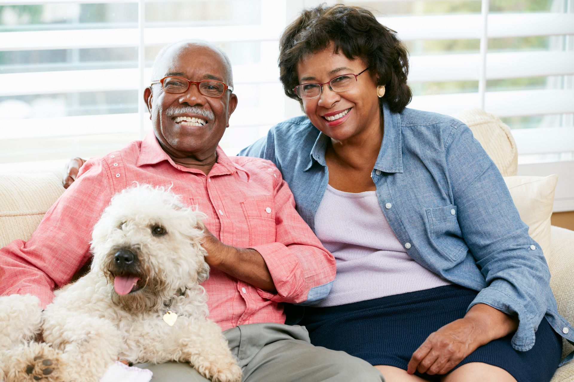Couple smiles with a fluffy dog on a couch, indoors. Man in red shirt, woman in blue.