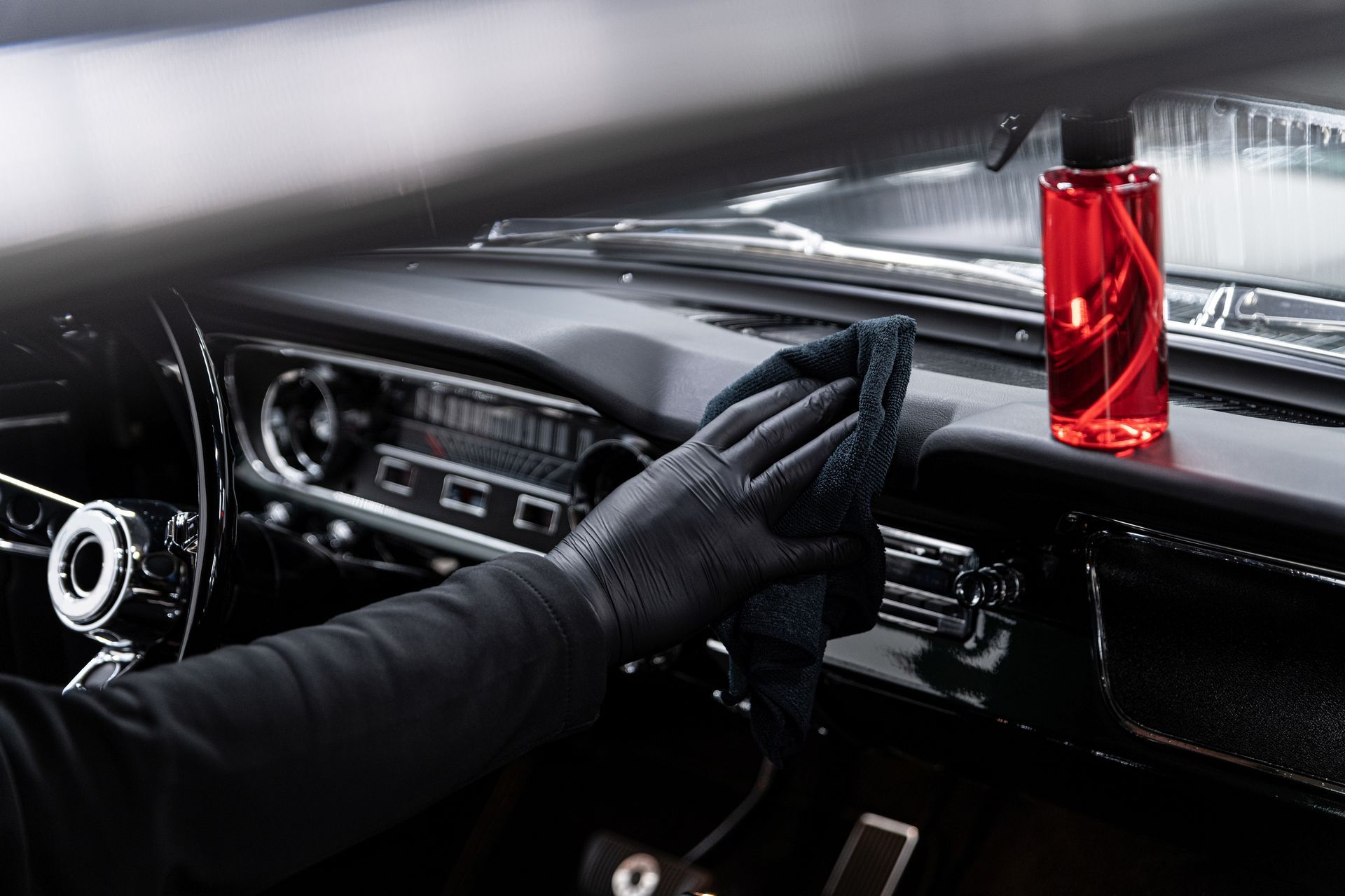 Black-gloved hand wiping a black car dashboard with a microfiber cloth; red spray bottle sits nearby.