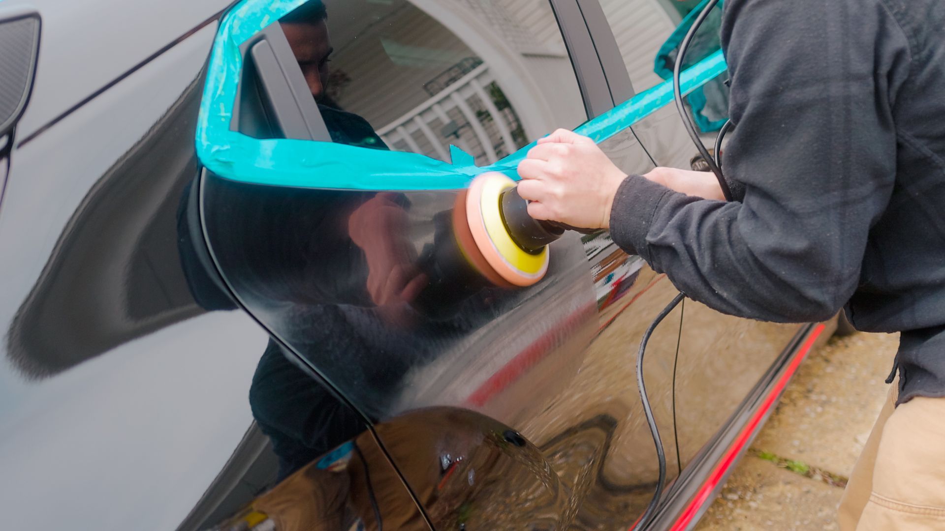 Person polishing a black car door with a buffing machine, outside.