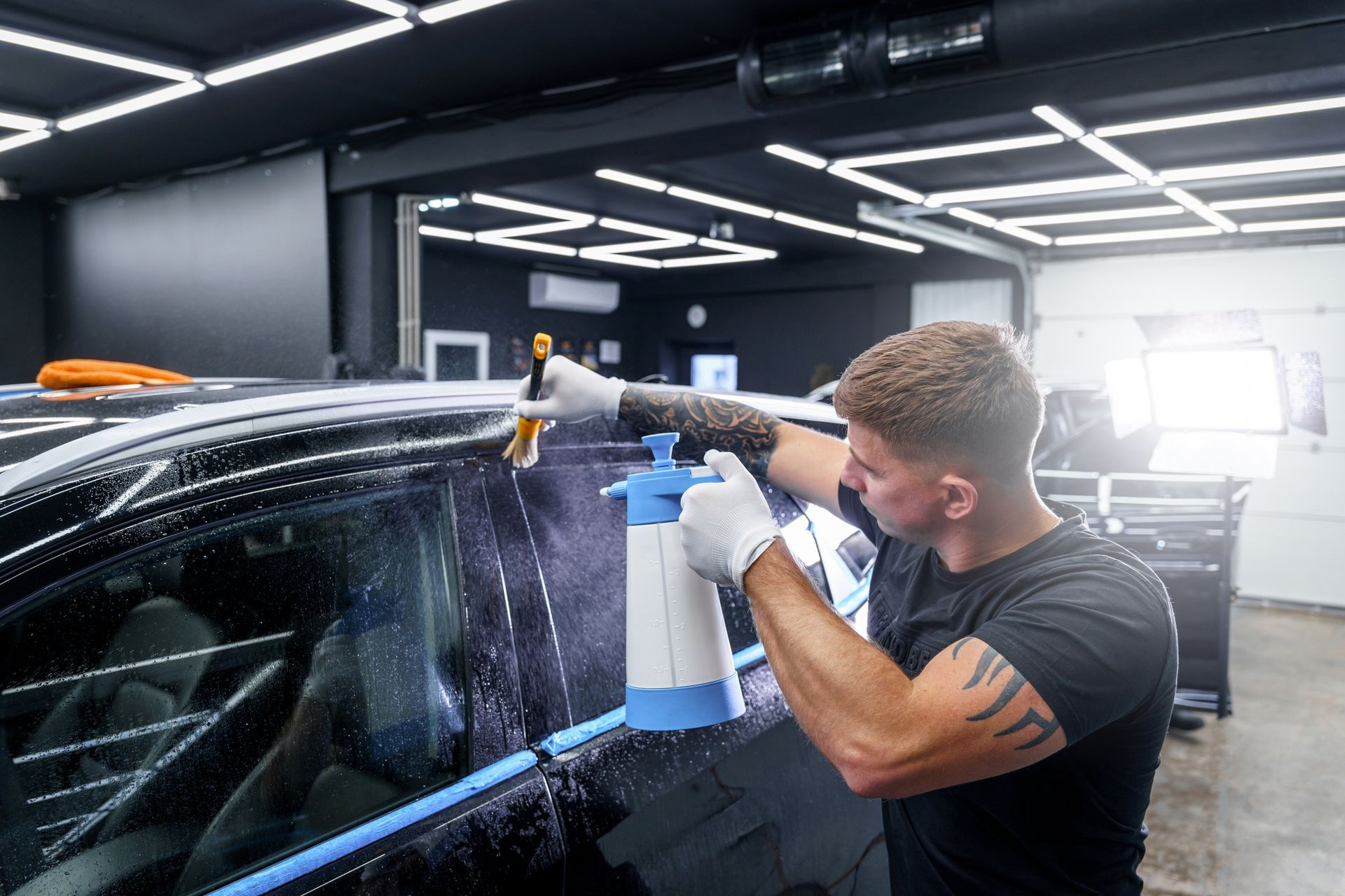 Man spraying soapy water on a car window in a garage, preparing to install tint.