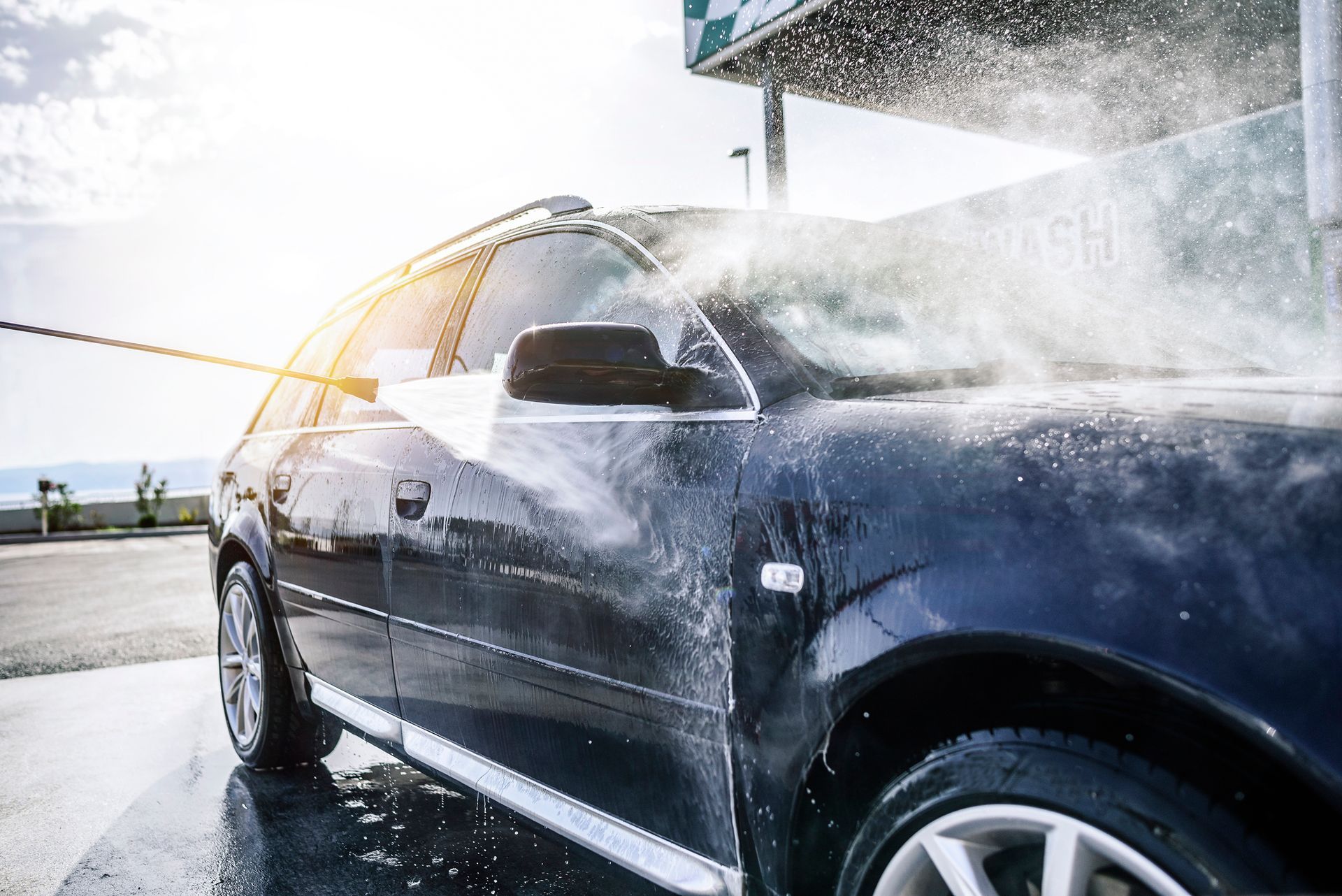 Dark car being washed with a pressure washer at a car wash on a sunny day.