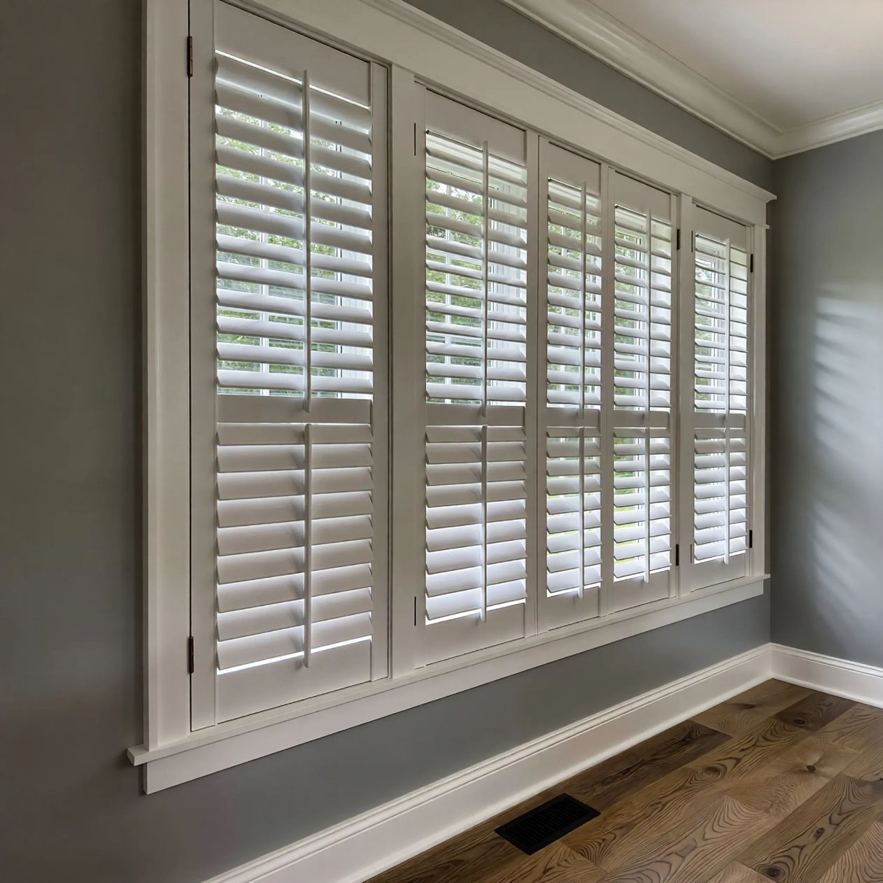 White plantation shutters installed on a multi-paneled window in a room with gray walls and wood flooring.
