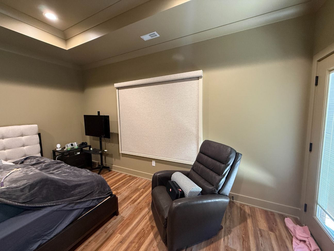 Bedroom featuring a grey armchair, a bed with grey bedding, a television on a stand, and a large window with white blinds.