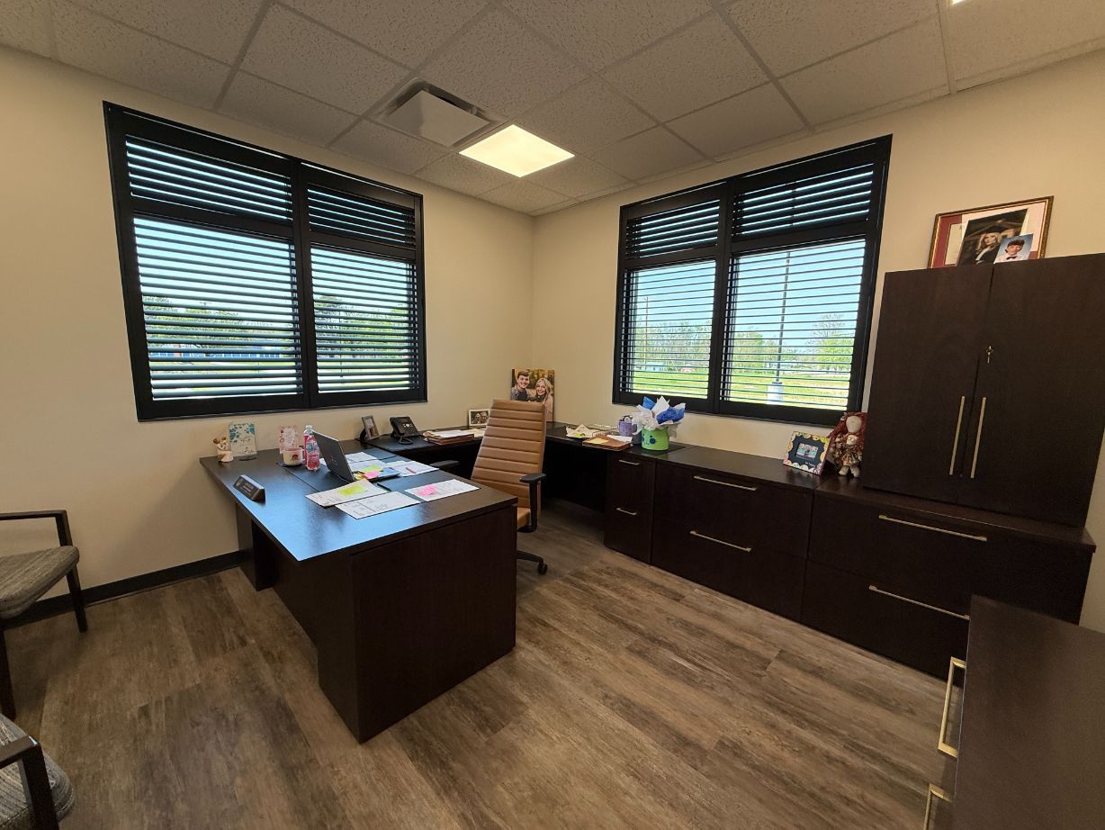 Modern office with two desks, dark cabinets, wood flooring, and large windows with shutters.