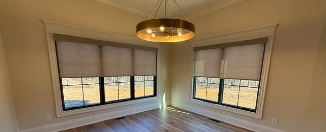 Empty corner room with large windows, beige roller shades, wood floors, and a brass ceiling light.