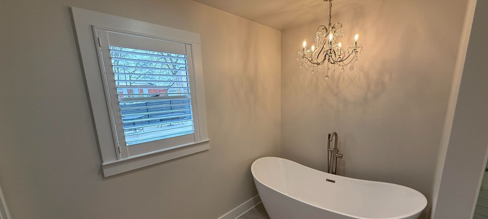 Bright bathroom with a white bathtub, chandelier, and window with plantation shutters.