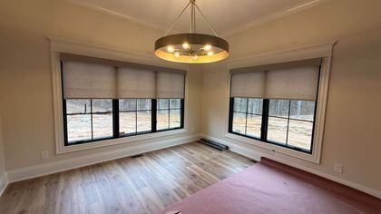 Empty room with two windows, wood floor, and chandelier; tan blinds and walls; windows have black frames.